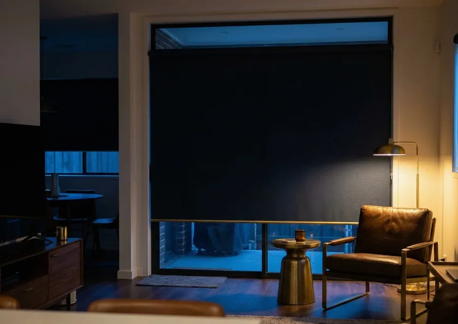 Blockout roller blind drawn fully down over a sliding door, holding a living room dark after sunset